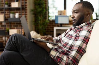 Sakallı siyah adam rahat oturma odasındaki kanepede çalışıyor, özgeçmişi inceliyor ve iş fırsatlarını araştırıyor. İş danışmanı uzak çalışma oturumu sırasında dizüstü bilgisayar kullanarak başlangıç belgelerini hazırlar.