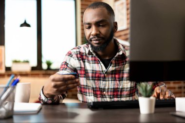 Çevrimiçi işlemleri yöneten Afrikalı Amerikalı girişimci, Merkez Ofis 'te kredi kartı bilgilerini okuyor. Uzaktan erkek yönetici tuğla duvar dairesindeki bilgisayarda finansal verileri güncelliyor.