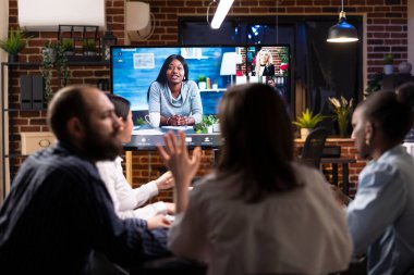 Çok ırklı meslektaşlar gece yarısı toplantıda proje planlarını tartışıyorlar. Uzak ekiple yapılan bir video konferansı, modern iş ortaklığını sembolize eden dijital ekranda görülebilir..