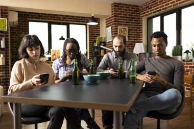 İçki ve abur cuburlarla masada oturan çok kültürlü arkadaş grubu mobil cihazlara odaklandı. Afrika kökenli Amerikalı ve Kafkasyalı bireyler sosyal medyada geziniyor ve cep telefonlarıyla mesajlaşıyor..