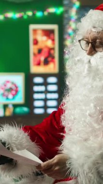 Dikey video. Mutlu adam, Noel karakterini canlandırıyor. Çocuklardan mektuplar okuyor, şapka ayarlıyor. Sahte sakal takmış, Noel Baba gibi gülümseyen, çocukların dilek listelerinin keyfini çıkaran, kamera A