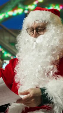 Dikey video Noel Baba çocuklardan gelen mektuplara bakar, onları selamlar ve karşılar. Noel Baba kostümlü gülümseyen kişi çocukların Noel için yazdığı dilek listesini okur, el sallar, selamlar.