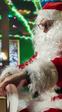 Dikey video Noel Baba hediye yapmak için ambalaj kağıdı kullanıyor. İyi çocuk listesini bitirdikten sonra. Hediye paketleyen, dünyayı gezmeye hazırlanan ve çocukları ve kamerayı ziyaret eden insanlar.