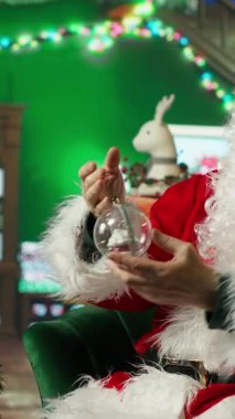 Dikey videolu profesyonel Noel Baba tripodla akıllı telefon ile çekim yapıyor, Noel ağacı kürelerini sunuyor ve hediyelerle, arka planlarla, çoraplarla, sahte şömineyle, süslemelerle ve diğer tatil malzemeleriyle hazırlanıyor.