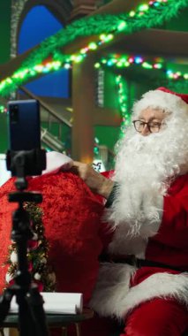 Noel Baba 'yı oynayan dikey videocu, cep telefonuyla tripodla çekim yapıyor, Noel arkaplanındaki çocuklara çorapları, şöminesi, süslemeleri ve diğer tatil süslerini göstermek için kırmızıdan hediyeler alıyor.
