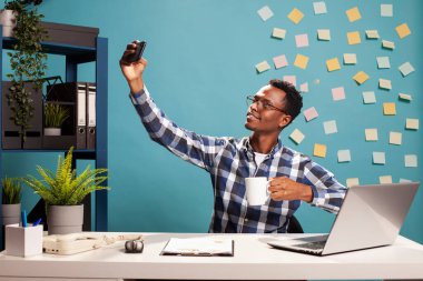 Elinde kahve olan siyahi bir muhasebeci ofis masasında oturmuş cep telefonuyla selfie çekiyor. Afrika kökenli Amerikalı yönetici elinde bir bardak ve cep telefonu ile iş günlerini kaydediyor..