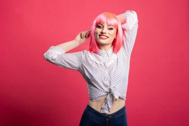 Güzel bir kadın, rahat ve esnek bir şekilde kameraya gülümsüyor. Gündelik giysiler içinde, kolları başının arkasında pembe arka planda poz veren neşeli genç bir kadın..