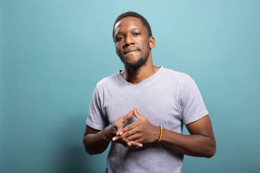 Sıradan tişörtlü Afro-Amerikan portresi, parmaklarını mavi arka plana yapıştırmış duruyor. Kendinden emin, büzüşmüş dudakları ve elleri bir arada kameraya bakan siyahi bir erkek..