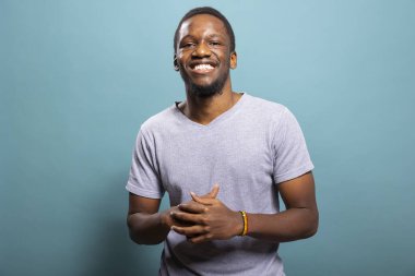 Genç siyahi adam sıkıca sarılmış elleri ve parlak bir gülümsemeyle kameraya bakarak poz veriyor. Afro-Amerikan erkek modelin portresi mavi arka planda duruyor, stüdyoda mutluluğu ifade ediyor..