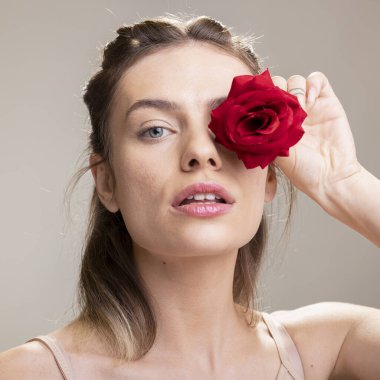 Bir güzellik reklamı için kırmızı çiçekle poz veren güzel bir cilt mankeni, sağlığın ve parlak taze cildin sembolü. Işıl ışıl, doğal görünüşlü, kendine güvenen genç bir kadın..