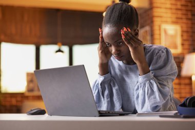 Serbest çalışan siyahi kadın, uzak çalışma programından tapınaklara masaj yaparak masaya yaslanıyor. Stresli girişimci dizüstü bilgisayara yorgun bir ifadeyle bakar, iş görevlerini evden yönetir.