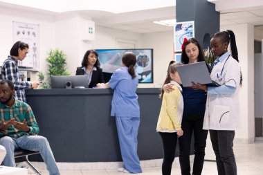 Hastane lobisinde çocuklu bir anneyle tıbbi profesyonel konuşmalar, dizüstü bilgisayardaki kontrol notlarını gözden geçirme. Klinik danışmanlığı sırasında anne ve kızına sağlık tavsiyesi ve teşhisi veren siyahi doktor.