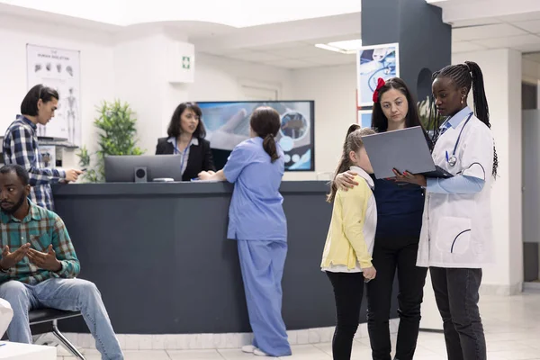 Hastane lobisinde çocuklu bir anneyle tıbbi profesyonel konuşmalar, dizüstü bilgisayardaki kontrol notlarını gözden geçirme. Klinik danışmanlığı sırasında anne ve kızına sağlık tavsiyesi ve teşhisi veren siyahi doktor.
