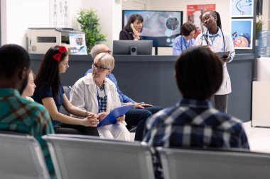 Doktor randevularından önce hastane lobisinde oturan çeşitli hastaların portresi. Klinik personeli ziyaretlere ve prosedürlere hazırlanırken, yaşlı kadın hekim sandalyedeki genç bayana destek veriyor