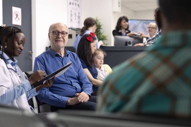 Smiling elderly man receives medical consultation in clinic waiting room. African american doctor with clipboard seated with old male patient in modern hospital lobby before appointment.