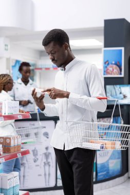 Black young adult searching pharmacy shop aisles and shelves, checking labels on prescription drugs for dosage information. Reading proper healthcare usage during pharmacy visit.