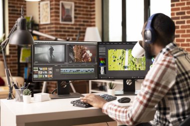Black freelance videographer sipping coffee while editing cinematic footage at home studio. Focused male editor works on transitions and color grading during post production session.
