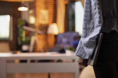 Female remote worker walking into apartment during sunset, laptop in hand. African American woman entrepreneur enters cozy living room, ready to focus on evening business projects.