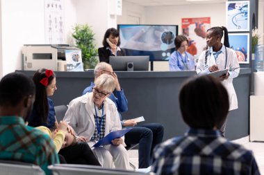 Busy hospital waiting room with elderly male doctor discussing treatment plans with mother and daughter. Female healthcare workers assist patients and manage medical records nearby.
