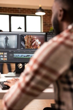 Masaüstü monitöründe film klipleri düzenleyen erkek video editörünün arka görünümü. Tuğla duvar dairesinden çalışan uzaktan kumandalı serbest çalışan, film sonrası sinemada sorunsuz bir içerik akışı sağlayan.