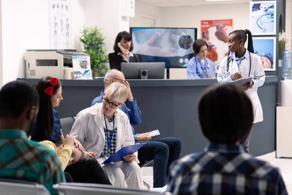 Busy hospital waiting room with elderly male doctor discussing treatment plans with mother and daughter. Female healthcare workers assist patients and manage medical records nearby.