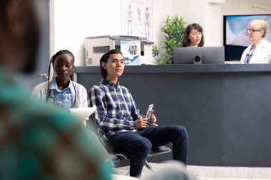 Young asian man seated beside female doctor in hospital lobby, waiting for consultation. Male clinic visitor holds promotional flyer, looking at camera and waiting in emergency room.