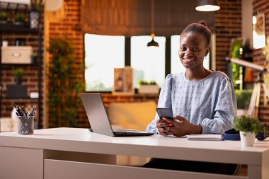 Neşeli siyah bayan girişimci dizüstü bilgisayar üzerinde çalışırken akıllı telefon kullanır. Genç kadın evdeki uzaktan çalışma ve dijital ağı dengeliyor, masada oturmuş telefon bekliyor..