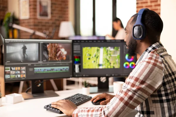 Profesyonel yazılım kullanarak kulaklıkla odaklanmış ses düzenleyicisi, görsel olarak ses parçalarını senkronize ediyor. İki monitörlü siyahi adam, ev stüdyosundaki ham kliplerden sinematik içerik hazırlıyor..