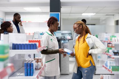 Black female pharmacist recommends painkillers to treat muscle pain, discussing with a patient dealing with a sore lower back. Woman with backache at the pharmacy shop. Back pain.