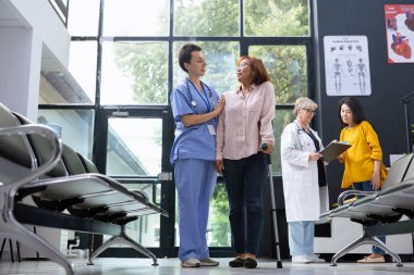 Hastane bekleme salonunda fizik tedavi sırasında koltuk değnekleriyle yürüyen Asyalı hastaya bakan hemşire. Sıhhiyeci kadın sorunlarına yardım ediyor, iyileşme süreci.