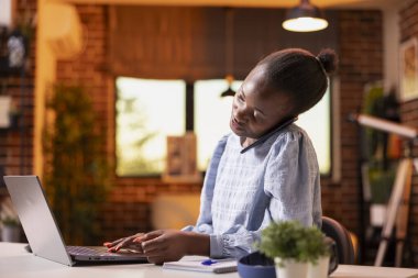 Siyahi kadın girişimci masada oturmuş, iş görüşmesi yapıyor ve dizüstü bilgisayardaki notları gözden geçiriyor, teknoloji ve iletişim becerilerini birleştiriyor. Evde cep telefonuyla konuşan Afrikalı Amerikalı kadın..