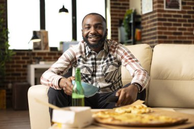 Tuğla duvar dairesindeki koltukta oturan mutlu adam elinde cipsler ve uzaktan kumandayla futbol maçı seyrediyor. Afro-amerikan kişi hafta sonu eğlencesini abur cuburlarla ve eve servis yemekleriyle geçiriyor.