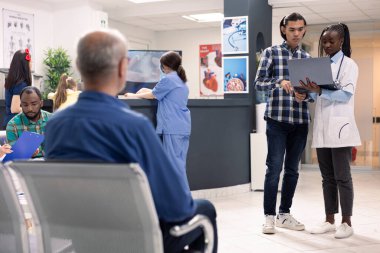 Laboratuvar önlüklü pratisyen doktor dizüstü bilgisayarla ayakta duruyor ve yoğun modern klinikteki hastalarla konuşuyor. Klinik ziyaretçileri acil serviste beklerken Asyalı adama dijital tıbbi sonuçlar gösteren kadın doktor.