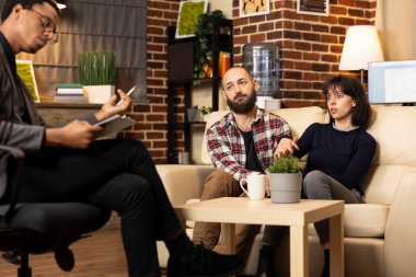 Endişeli beyaz kadın, psikoterapi seansında erkek arkadaşının yanında üzgün bir ifadeyle oturuyor. Afrika kökenli Amerikalı terapist, çiftle stres ve zihinsel refahı tartışıyor..