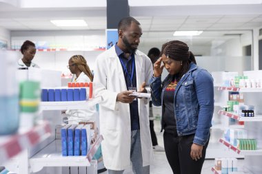 Pharmacist service scene where a professional gives headache drugs remedy, explains pharmaceutical treatment and medication dosage to a woman at a busy pharmacy dispensary.