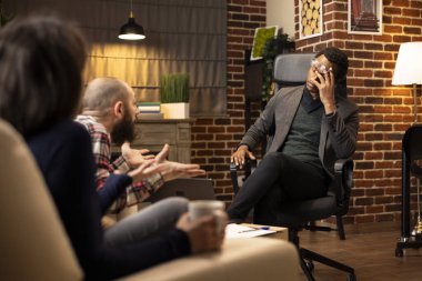 Black male psychologist rubs head in frustration during couple therapy session, showing stress while listening to caucasian man. Irritated African American therapist in modern brick wall office.