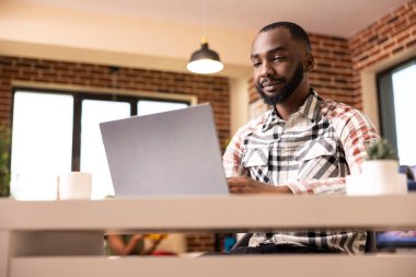Young african american man seated at table, updating schedule and editing online content on laptop. Remote male copywriter proofreading marketing materials in cozy home office.