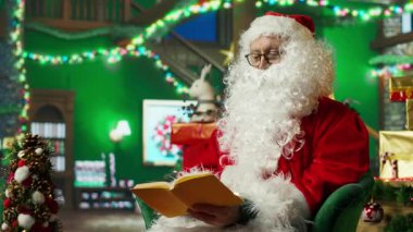 Santa Claus reading book in workshop after finishing list of kids who has been naughty or nice. Older person wearing fake beard enjoying novel at home before starting trip to visit children, camera A