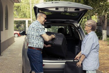 Cheerful elderly people prepare for a summer trip and organizing baggage in the car trunk. Joyful retirement lifestyle with road trip exploration and outdoor fun with life partner.