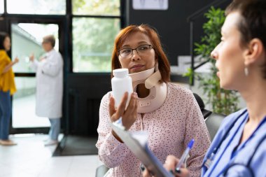 Hastane ziyareti sırasında boyun travması geçiren hasta omurilik desteği takıyor. Doktor acı veren semptomları ve iyileşme sürecini hafifletmek için ilaç ve fizyoterapi öneriyor..