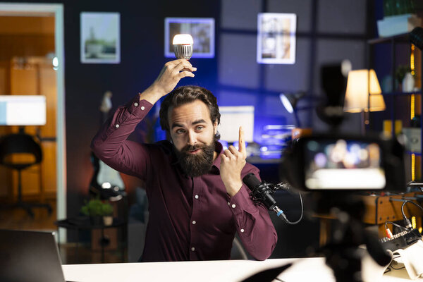 Man using light bulb to illustrate brilliant idea, filming with professional camera. Happy media star pointing finger up to show inspiration concept, recording with specialized gear
