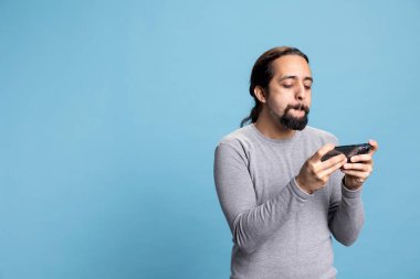 Erkek oyuncu akıllı telefonunda mobil video oyunlarıyla eğleniyor, mavi arka planda internet sporları turnuvasının tadını çıkarıyor. Genç yetişkin kameranın önünde siber oyun oynuyor..