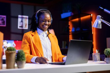 Happy woman wearing headphones laughing, enjoying watching comedy TV show during relaxation time at home. Cheerful person sitting at desk amused by funny television series on laptop
