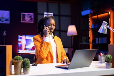 African american teleworker receiving phone call from job colleague while working from home on laptop. Freelancer speaking with coworker using smartphone while solving tasks