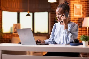 Black woman working from home, using smartphone for business calls and checking client updates on laptop. Female freelancer multitasks with phone and digital device in cozy living room.