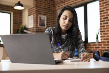 Hintli kadın profesyonel, tuğla duvar dairesinde dizüstü bilgisayarın yanında notlar alıyor. Merkez ofisteki not defterine stratejik iş planlarını özetleyen konsantre iş kadını..