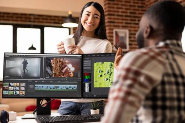 Young asian woman cheerfully sharing coffee break with african american boyfriend during home editing session. Happy couple enjoying relaxed conversation at workstation setup in brick wall living room