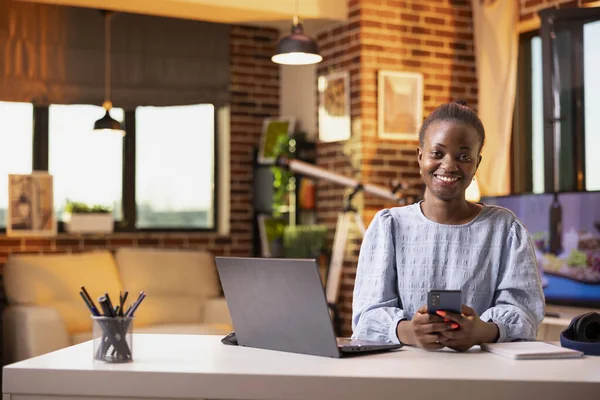 Portrait of young businesswoman using cellphone and looking at camera. Self employed woman sits at table with laptop, networking online and replying to messages on mobile device.