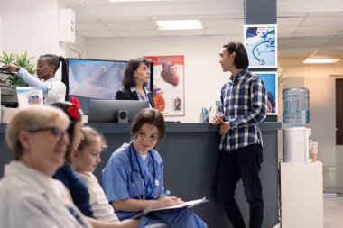 Receptionist at hospital front desk, talks to asian man and manages his doctor appointment on computer. Medical administrative assistant registers patient and provides healthcare information in clinic