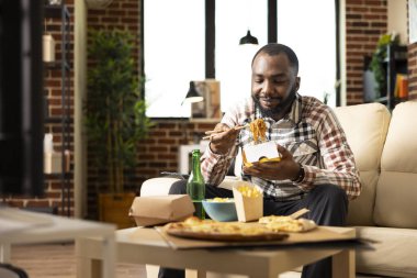 Afro-Amerikalı bir adam rahat bir şekilde kanepede oturup evde film izlerken lezzetli erişteler yiyor. Genç bir erkek, oturma odasında yemek yiyor ve televizyon seyrediyor..